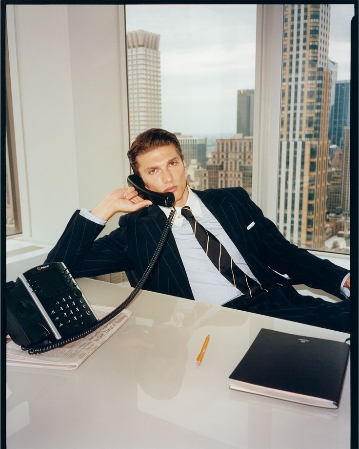 A founder in a pinstripe suit leans back at a desk in a high-rise office, taking a call on a desk phone, with a city skyline through the window behind him.